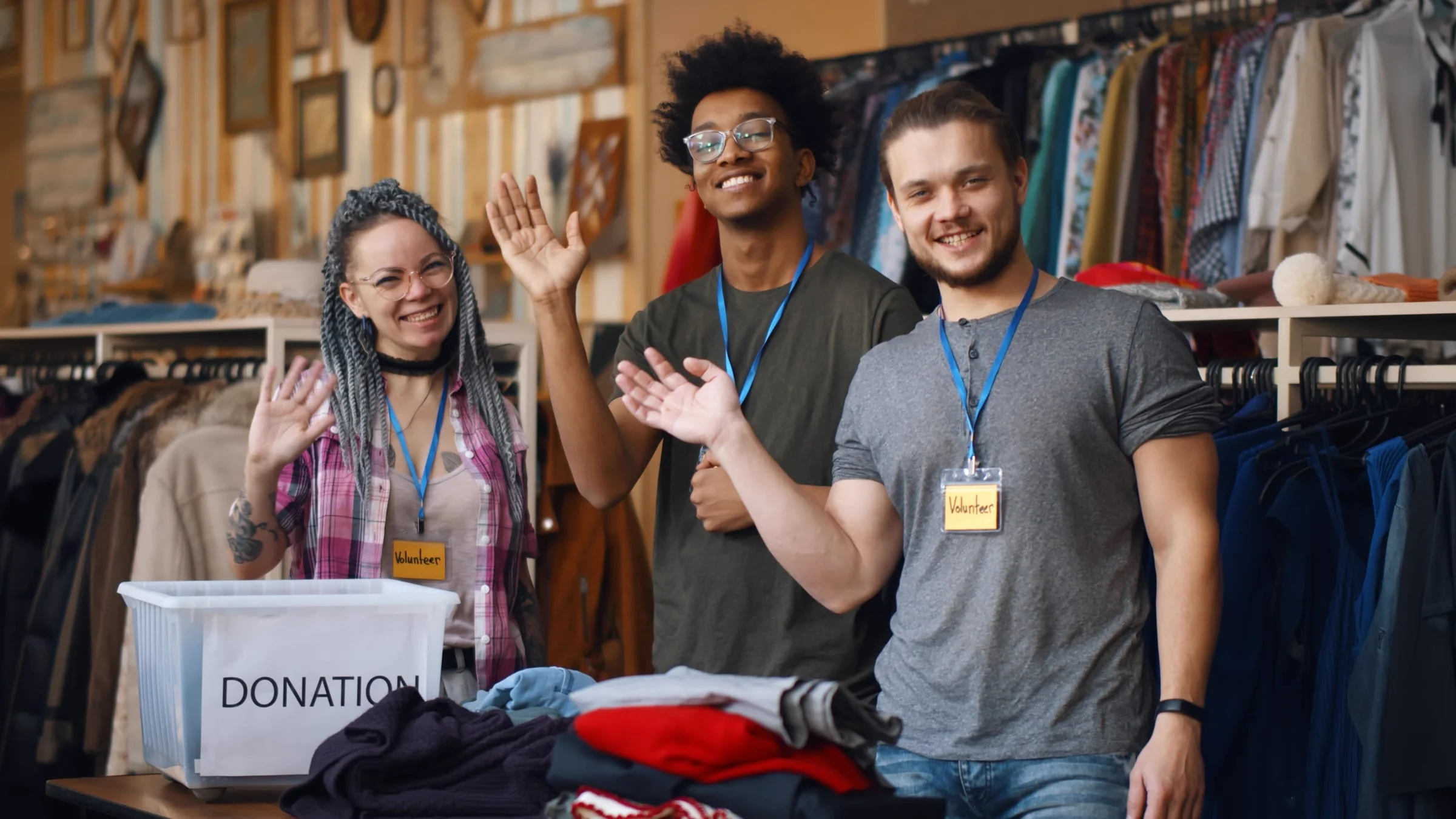 Three volunteers waving beside racks of donated clothing